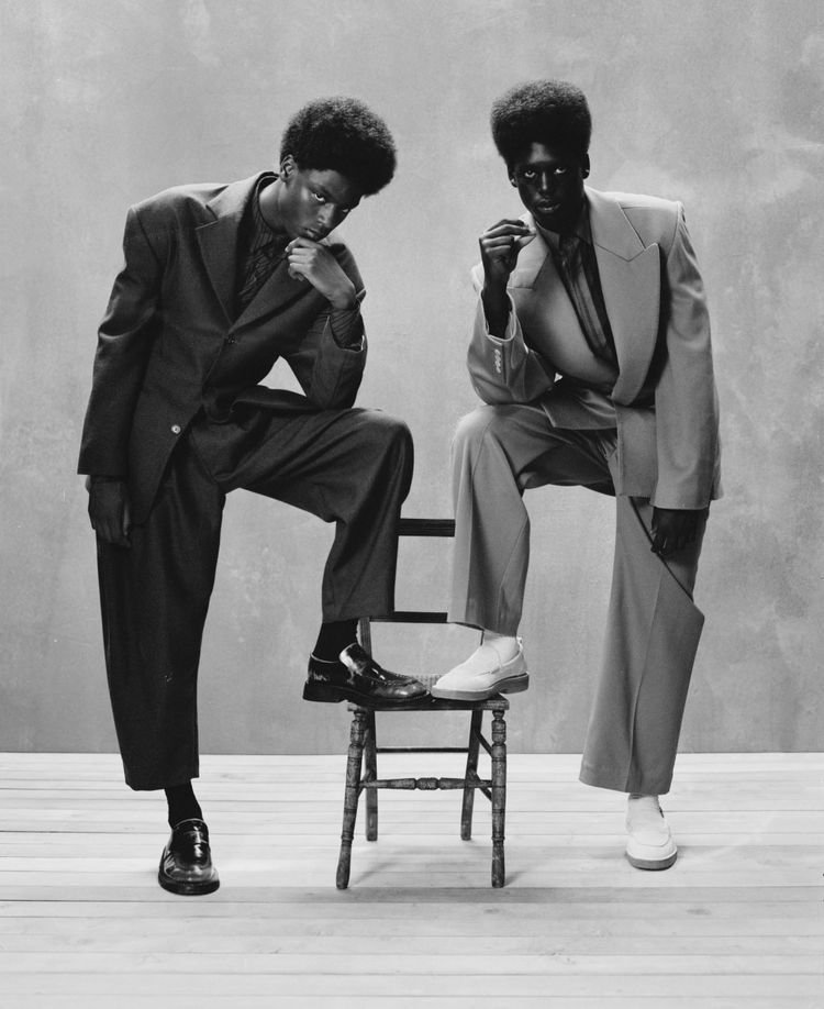 Two models in oversized tailoring around a wooden chair, black and white