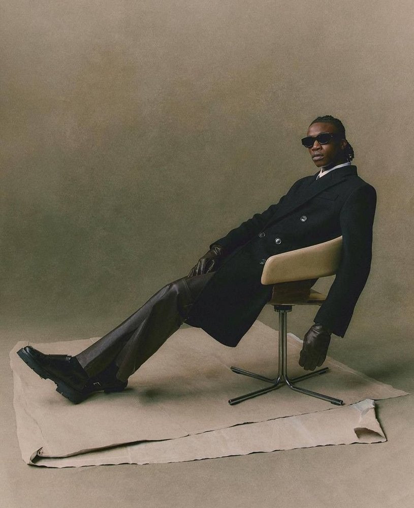 Model in sunglasses, a long dark coat and leather trousers reclining on a chair on a canvas backdrop