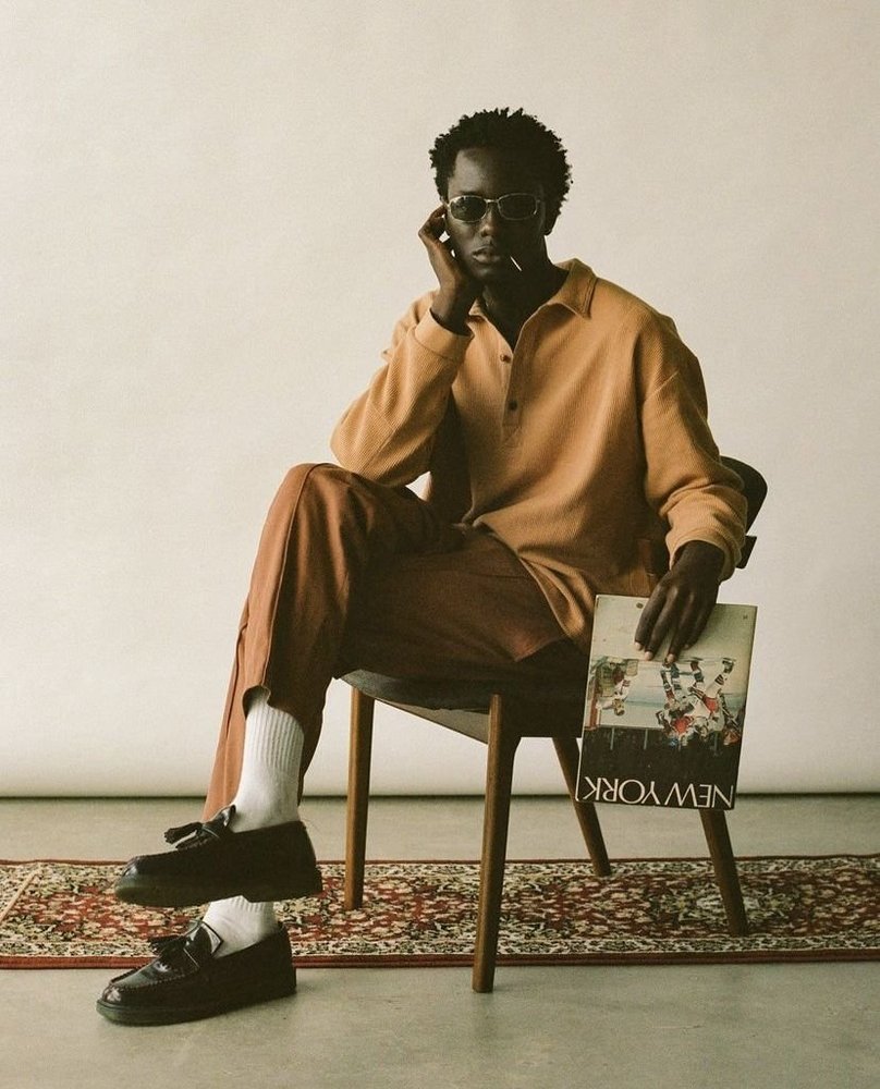 Model in a rust knit polo and trousers with white socks and loafers, seated on a wooden chair on a rug