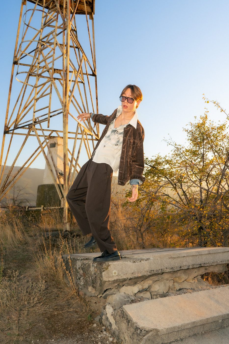 Tomislav Ordanov in tinted glasses, a brown velvet jacket, printed shirt and wide brown trousers, posing on a concrete ledge beside a steel tower at sunset